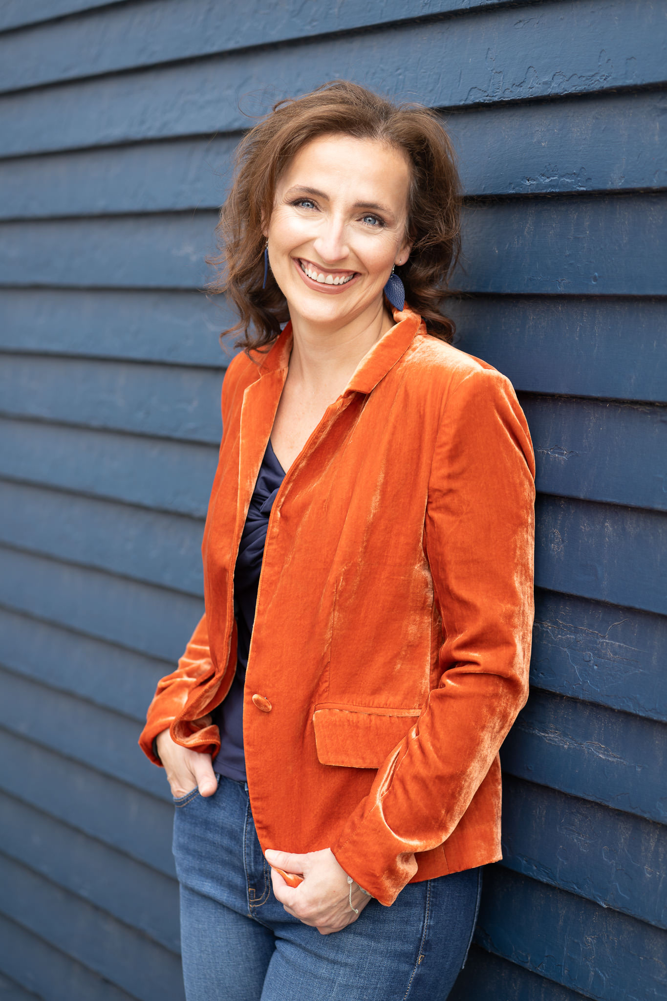 Erin smiling while leaning against blue siding in a burnt orange velvet blazer.